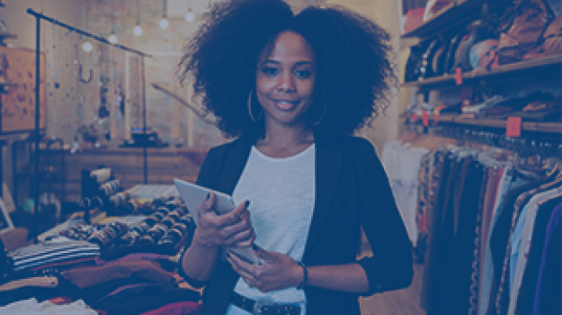 A Black, female business owner stands in her clothing shop while holding a tablet.
